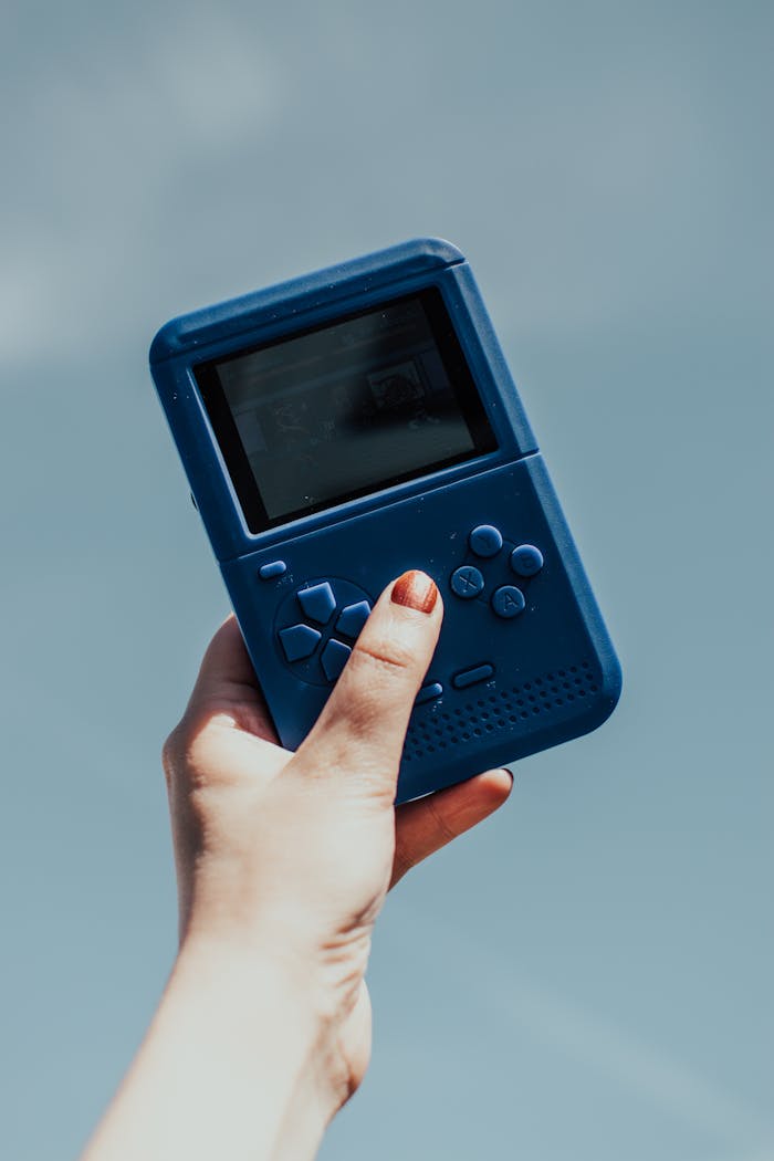 our-services-1 A close-up of a hand holding a vintage blue gaming console against a clear sky.